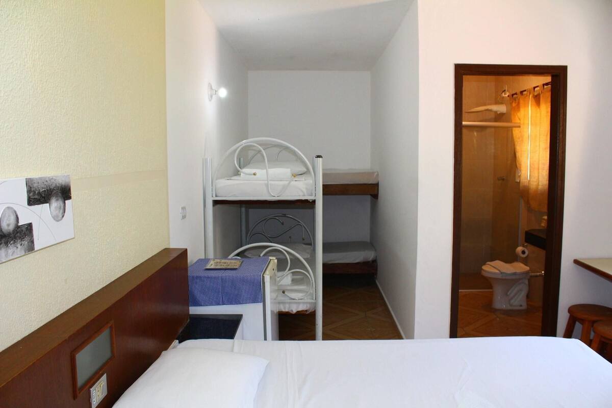 Quarto de hotel pequeno e compacto, com uma cama de solteiro, uma mesinha e uma beliche.