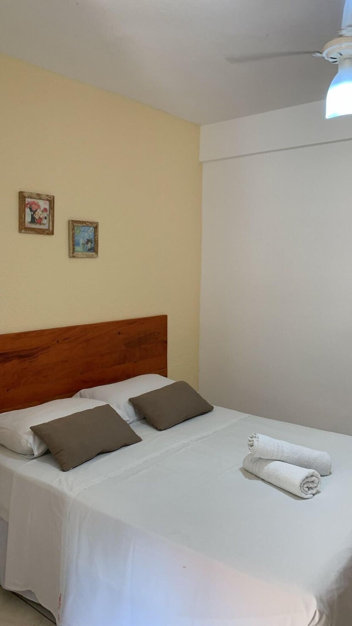 A neatly made bed with white linens and two brown pillows, a wooden headboard, and two framed pictures on the wall.