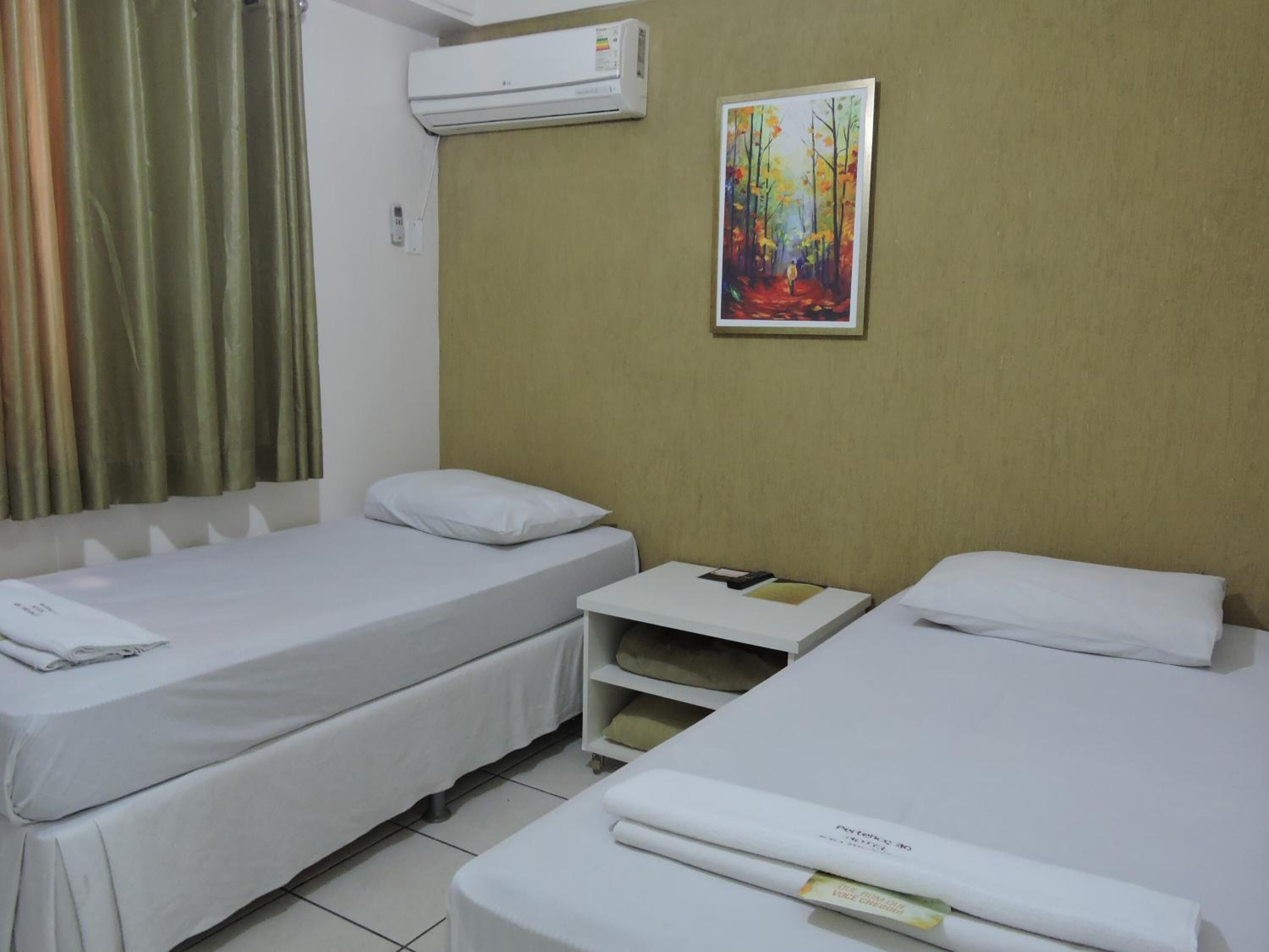 Quarto de hotel com duas camas de solteiro, uma mesinha, ar condicionado e um quadro na parede.