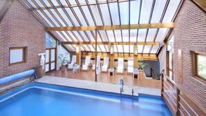 A swimming pool with a wooden deck and lounge chairs, surrounded by brick walls and large windows.