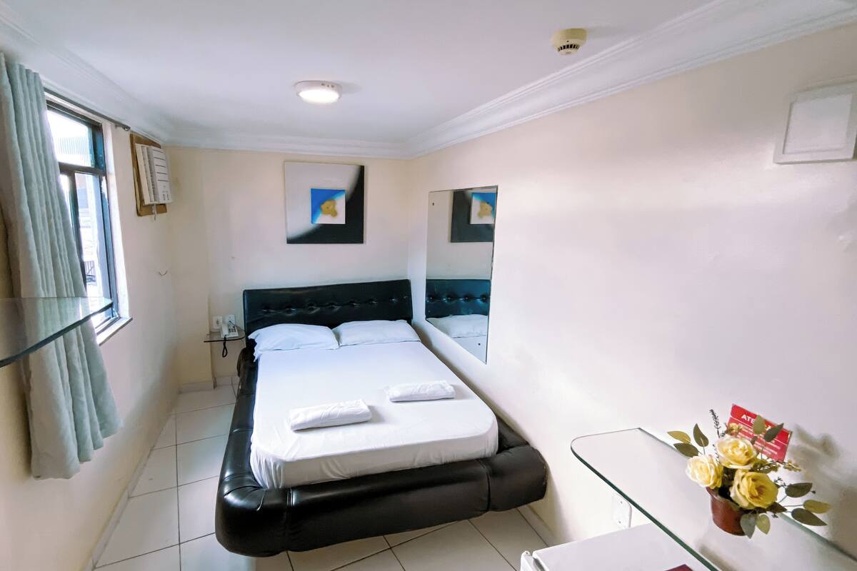 A hotel room with a bed, a bedside table, a window with curtains, a wall-mounted air conditioner, and a mirror.