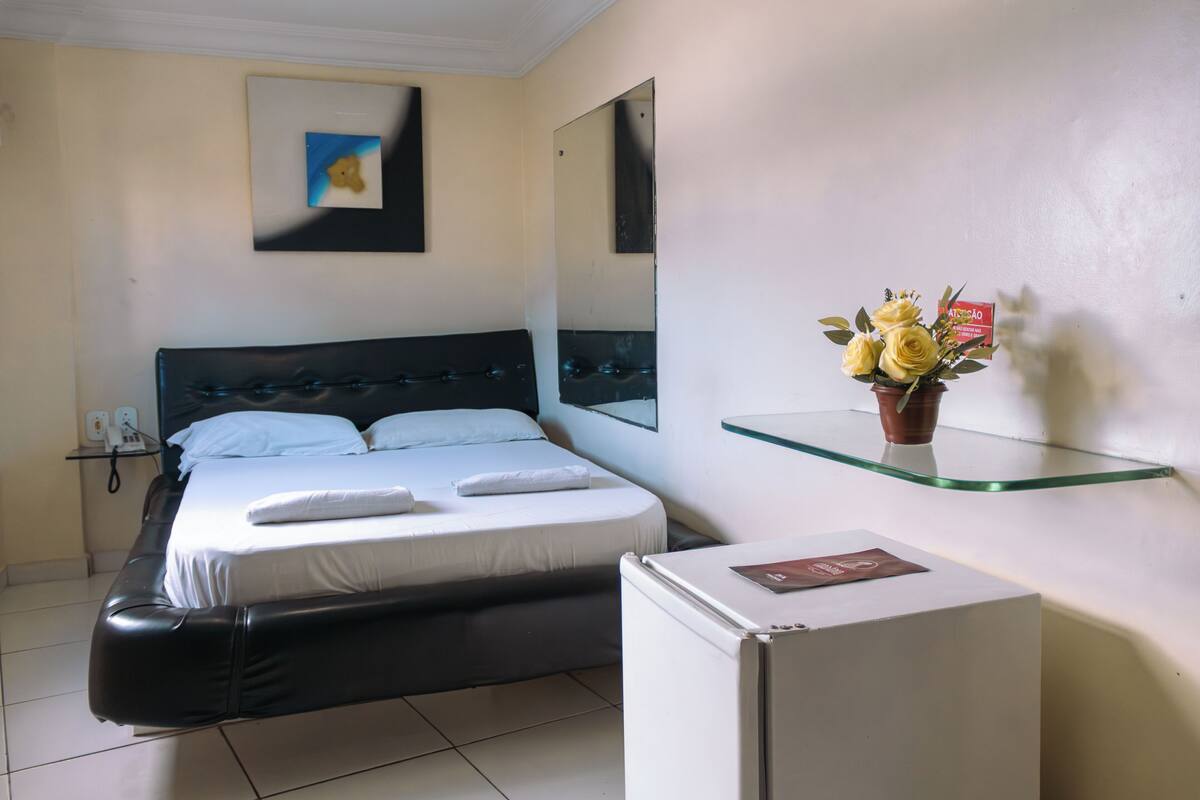 A hotel room with a bed, a small refrigerator, a bedside table, a mirror, a wall-mounted shelf with a potted plant, and a framed picture.