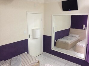 A small, compact room with a single bed, a bedside table, a mirror, and a television mounted on the wall. - Hotel Orquídea (São Bernardo do Campo)