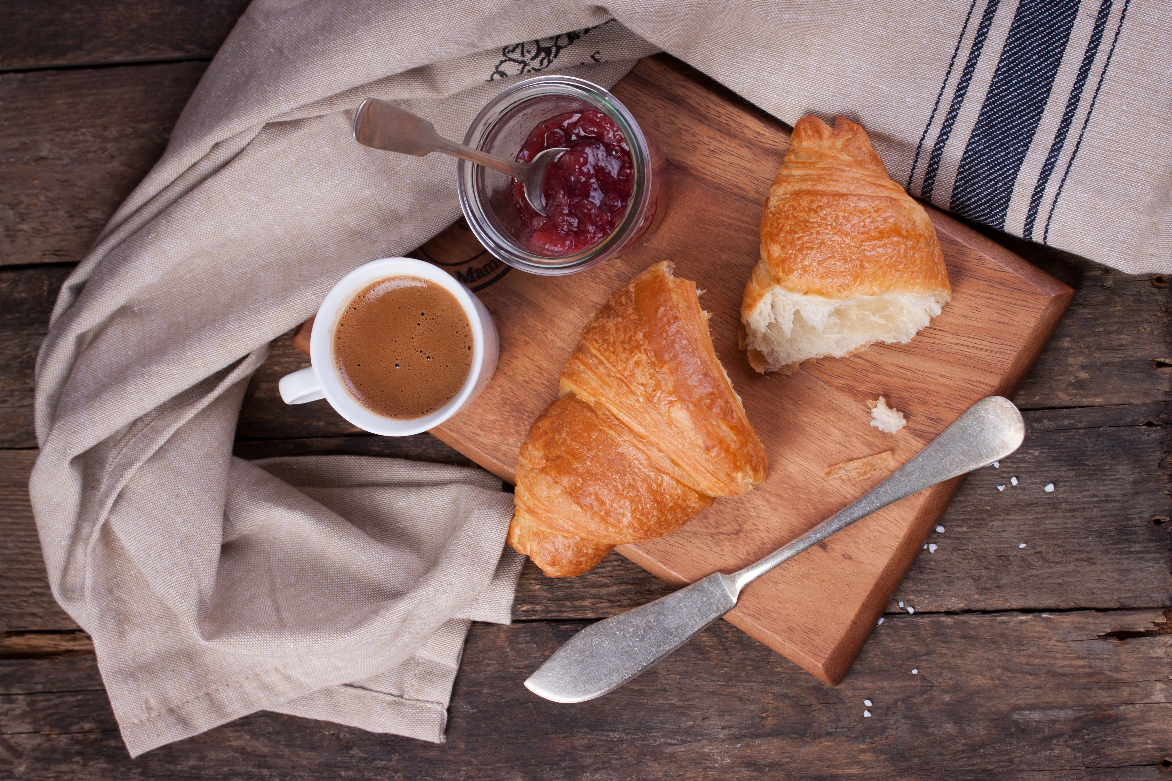 A wooden table with a croissant, a jar of jam, and a cup of coffee.