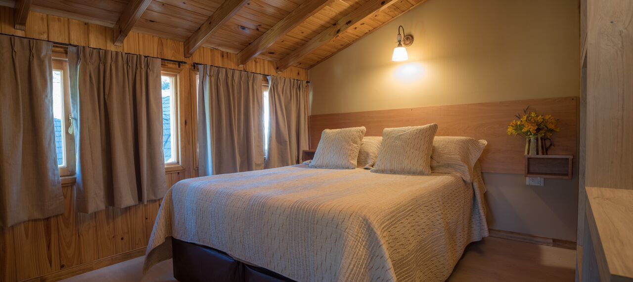 Un dormitorio acogedor con techo de madera, una cama grande con manta color beige y una mesita de noche con un florero.