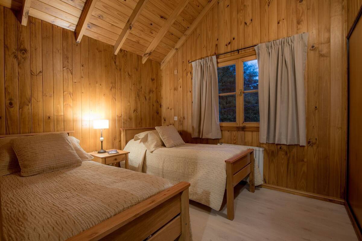 A wooden cabin bedroom with two beds, a nightstand, a lamp, and a window with curtains.