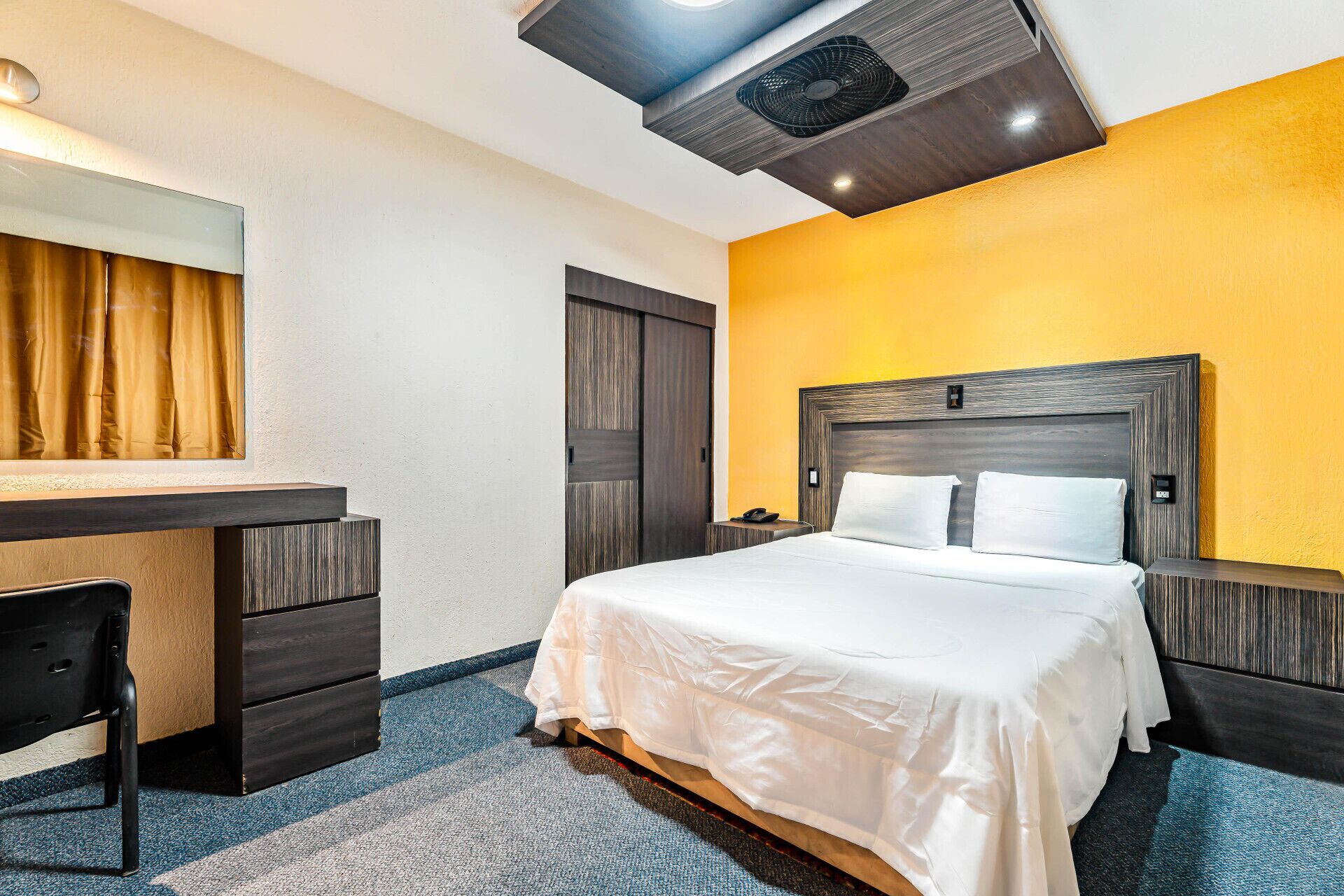 Una habitación de hotel con una cama, un escritorio, una silla, un espejo y un ventilador de techo.