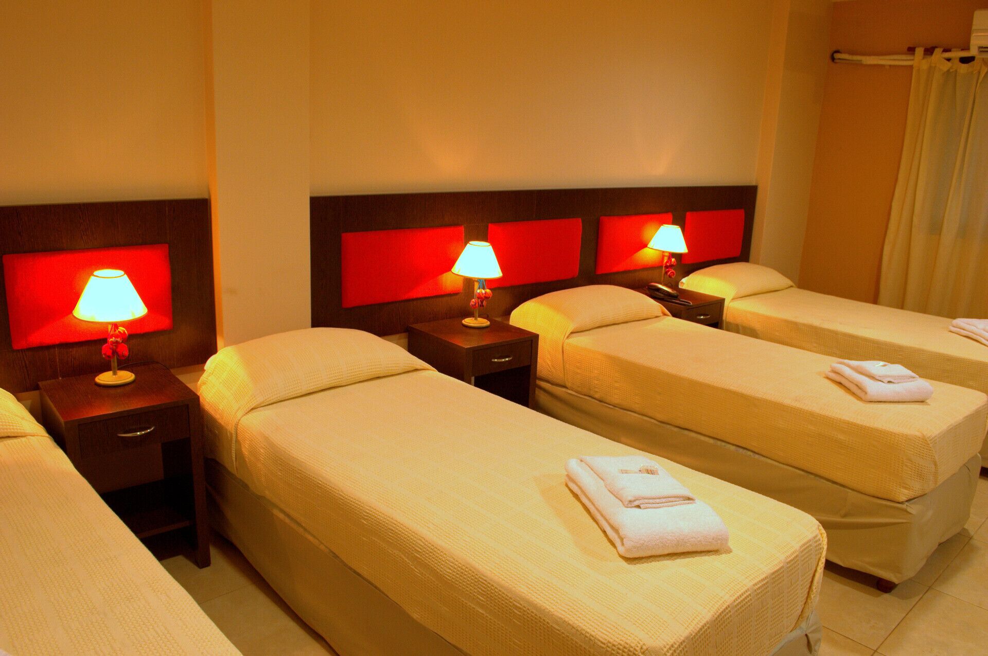 Ein Hotelzimmer mit drei Einzelbetten, jeweils mit Nachttisch und Lampe. Das Zimmer hat eine warme Farbgebung mit roten Akzenten an den Kopfteilen und einem Vorhang am Fenster.