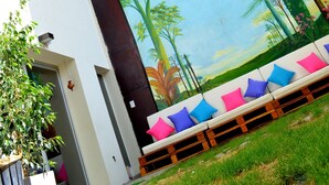 A modern building with a mural of a forest scene on the lower wall, a white exterior, and a wooden bench with colorful cushions. - Jaque Mate Hostel (Mendoza)