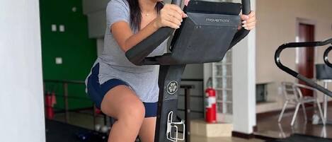 A woman using an exercise bike in a gym with a green wall, a treadmill, and a fire extinguisher in the background.