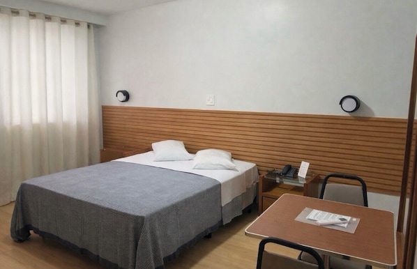 Quarto de hotel com cama, mesa, cadeira e criado-mudo com telefone.