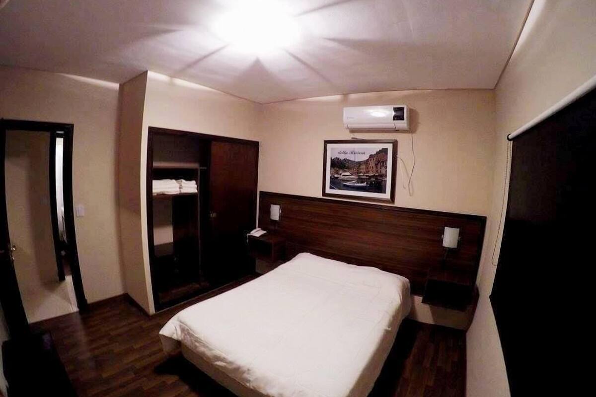 A hotel room with a bed, a wardrobe, a bedside table, an air conditioning unit, and a framed picture on the wall.