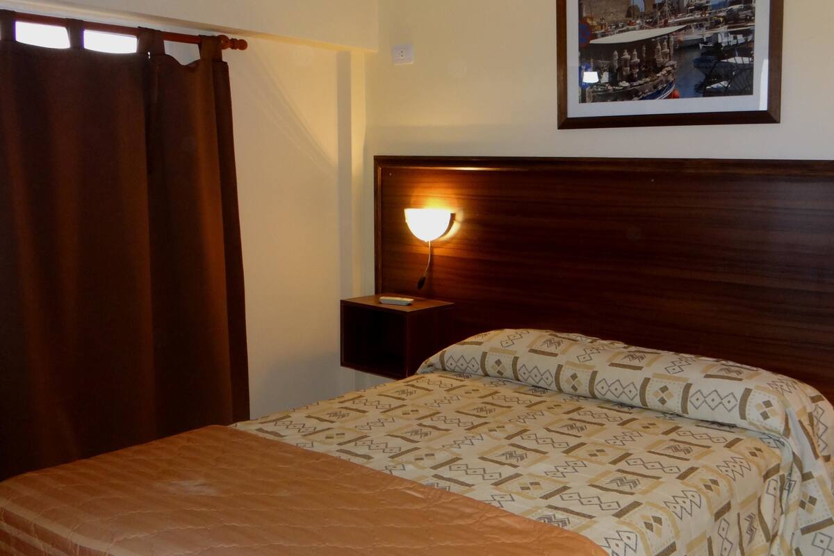 A hotel room with a bed, a nightstand, a lamp, a framed picture of a coastal scene, and a window with curtains.