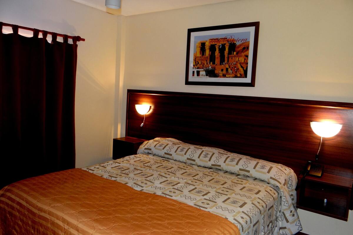 A hotel room with a large bed, two bedside lamps, a framed picture on the wall, and a dark curtain.