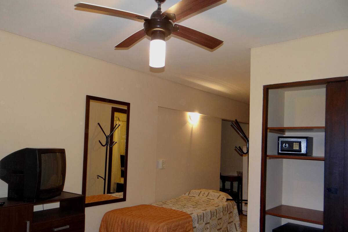 A bedroom with a ceiling fan, a bed with a patterned bedspread, a dresser with a television on top, and a mirror on the wall.