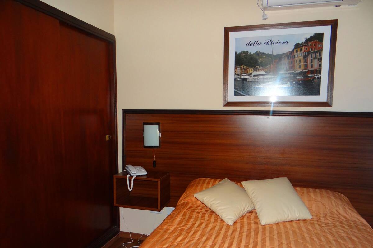 A hotel room with a wooden headboard, a bed with striped bedding, two pillows, a bedside table with a phone, and a framed picture of a cityscape on the wall.
