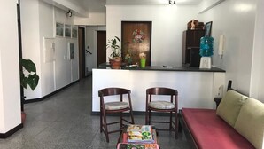 A small, well-lit room with a kitchenette, two wooden chairs, a small table with children's books, a red sofa, and a water dispenser.