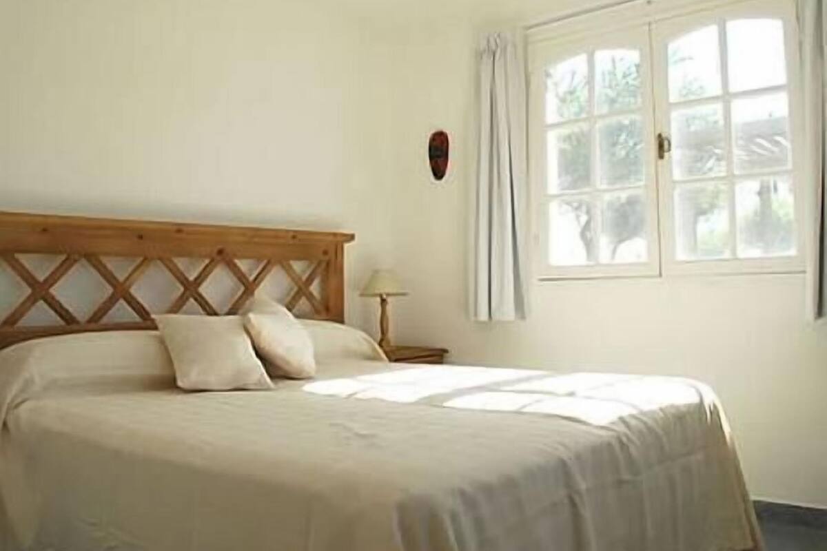 A neatly made bed with a wooden headboard in a room with a window and a nightstand with a lamp.