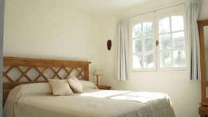 Un dormitorio con cabecera de madera, cama con ropa de cama blanca, mesita de noche con lámpara y ventana con cortinas.