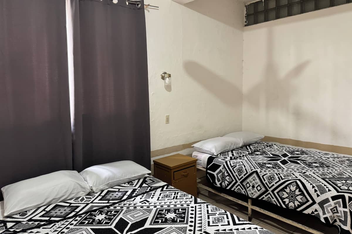 Una habitación pequeña y sencilla con dos camas, una mesita de noche, una lámpara en la pared y un ventilador de techo.