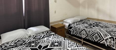 A small, simple room with two beds, a nightstand, a wall-mounted light, and a ceiling fan.