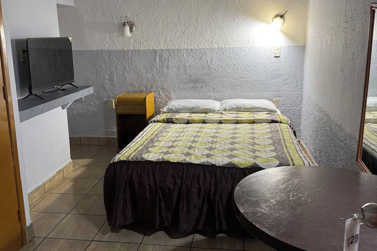 Una habitación con cama, una mesita, un televisor sobre un mueble y una lámpara fijada a la pared.