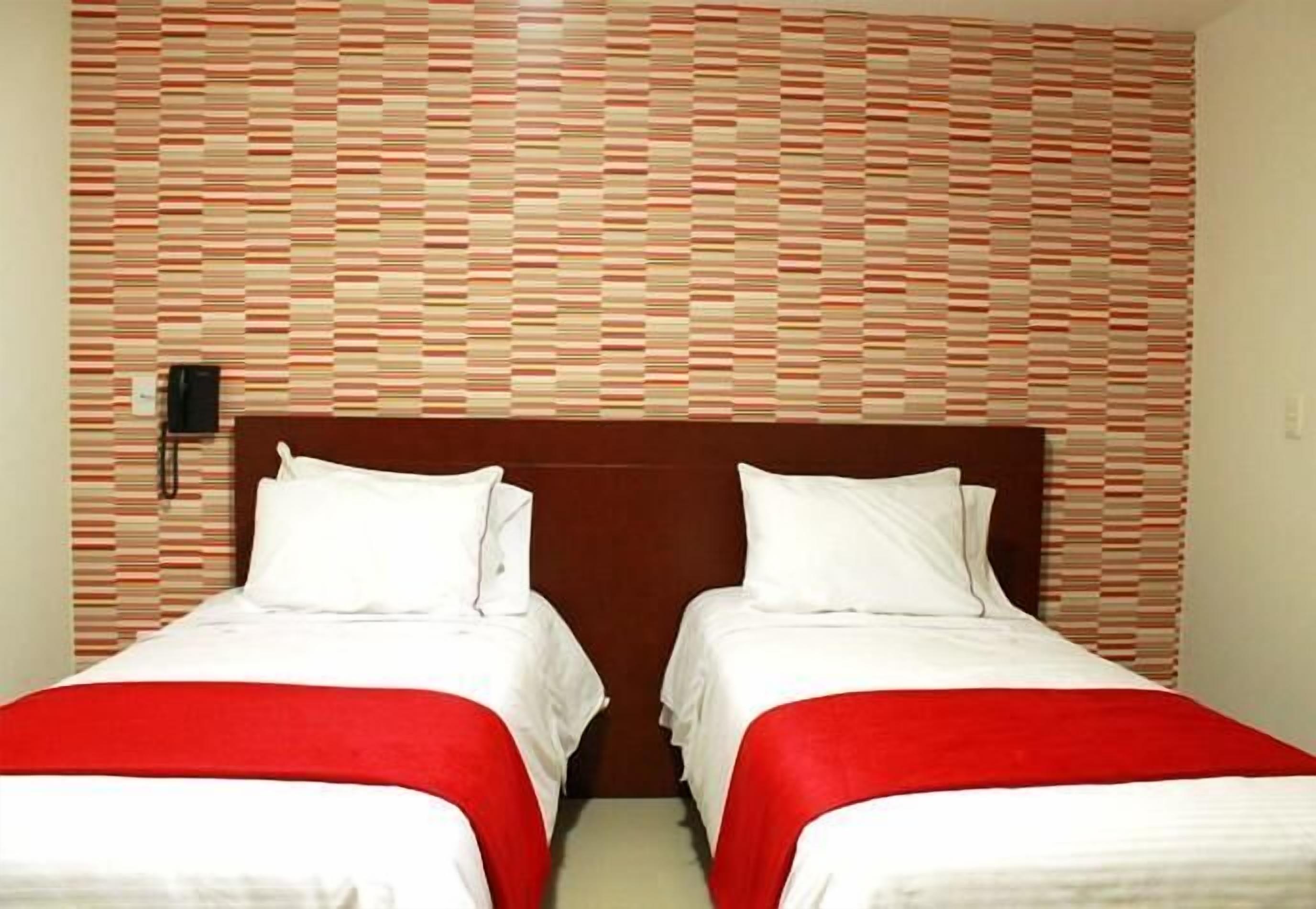 A hotel room with two beds, each with white bedding and red throw blankets. The room features a patterned wall and a wooden headboard.