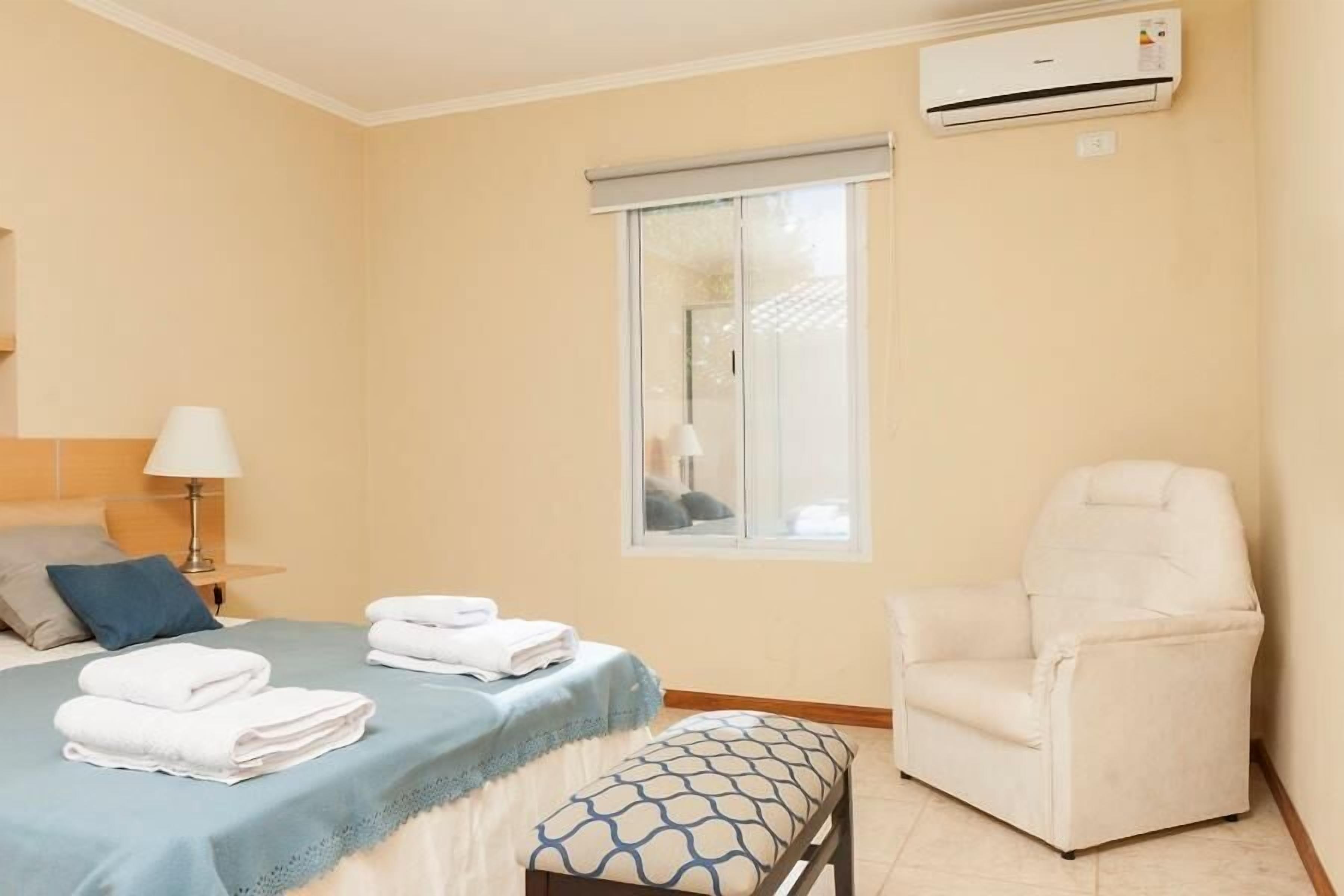 Una habitación de hotel con una cama, una silla, un banco, un ventanal y un equipo de aire acondicionado.