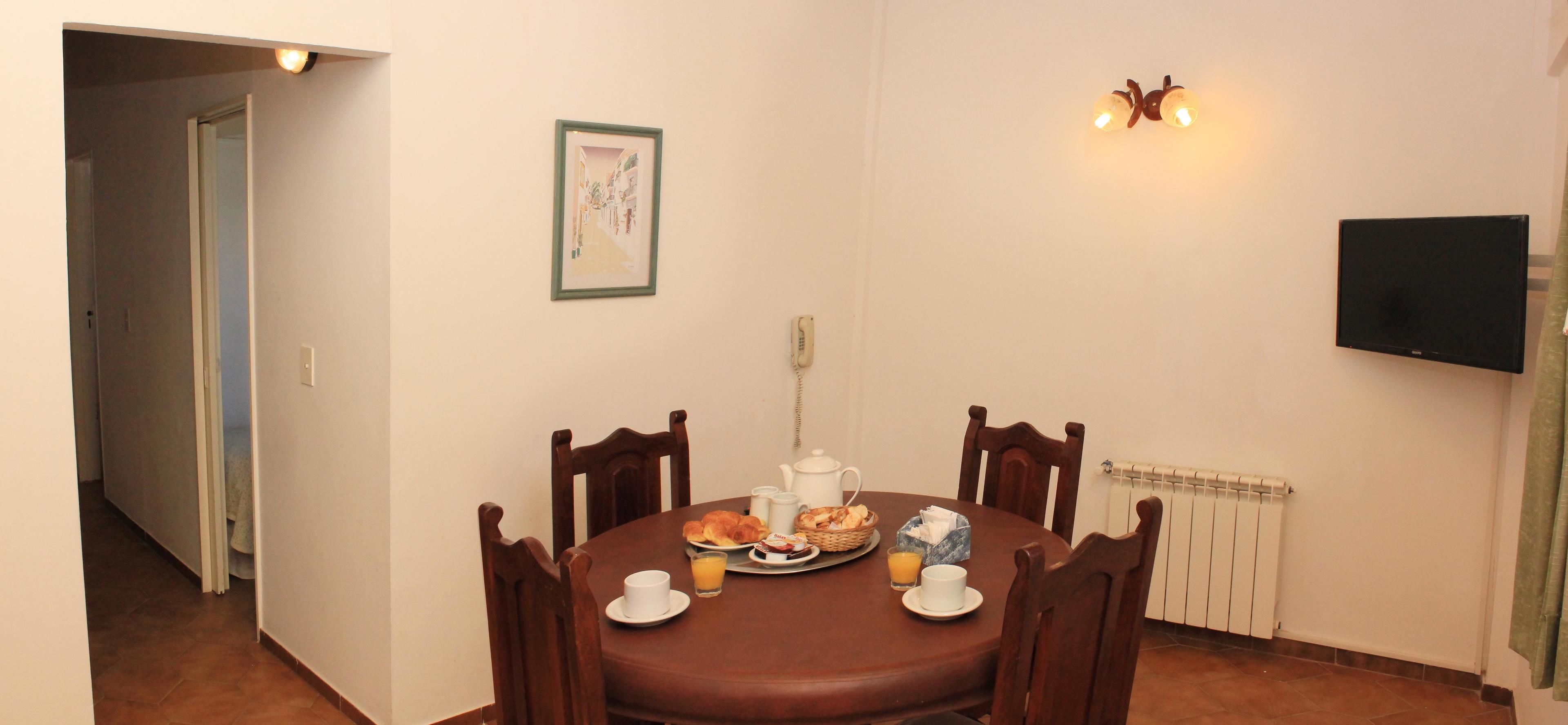 A dining area with a round table, chairs, breakfast items, a painting, a telephone, and a TV on the wall.