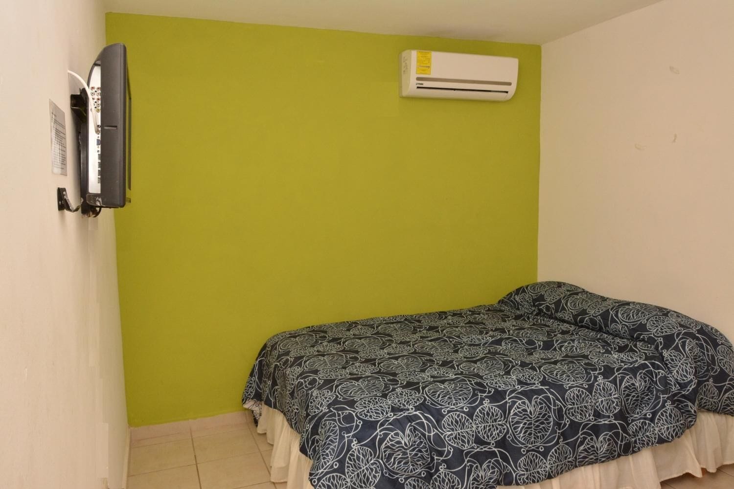A small room with a bed, an air conditioner, and a wall-mounted television.