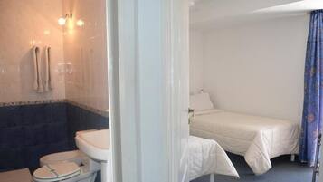 Habitación de hotel con baño que incluye inodoro, lavamanos y ducha. Cuenta con una cama con cubrecama blanco y una cortina azul.