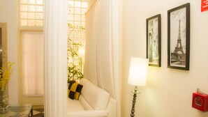 Habitación de hotel con sofá blanco, mesa de centro de vidrio, lámpara, un jarrón con flores, ventana con cortinas y dos cuadros con imágenes de la Torre Eiffel.