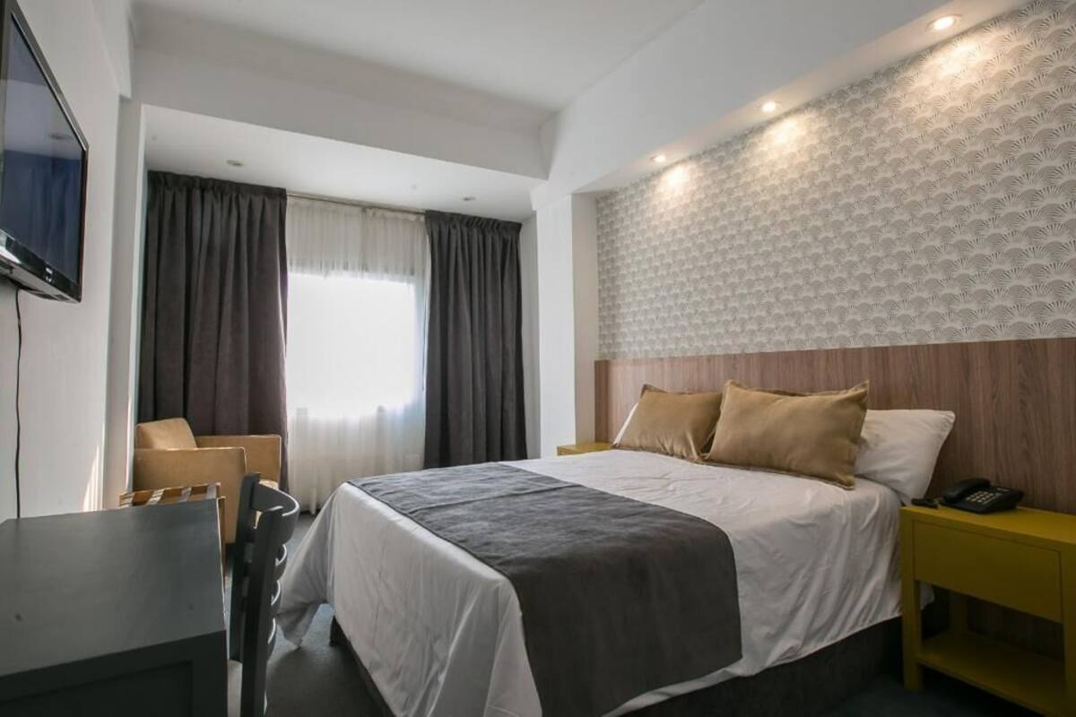 A hotel room with a bed, a desk, a chair, a TV, and a window with curtains.