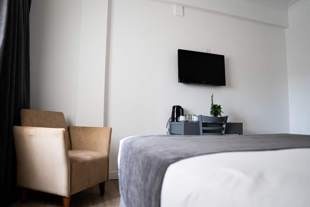 A hotel room with a bed, a chair, a TV mounted on the wall, and a small table with a plant and a coffee maker.