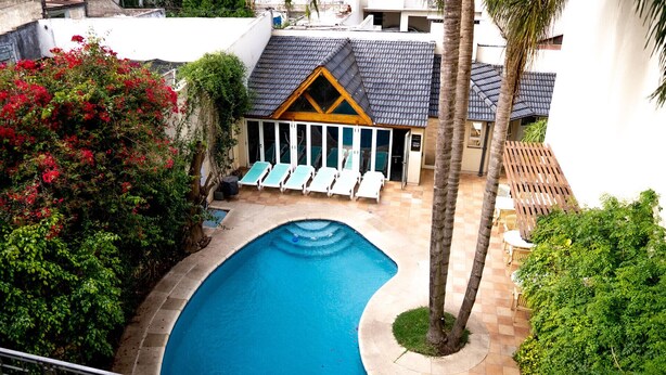 Une piscine à bordures courbes, entourée de chaises longues et d’un petit bâtiment doté d’un toit en pente.