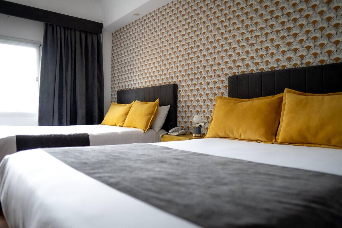 A hotel room with two beds, each with a black headboard and white bedding. The room features a patterned wallpaper and a large window with dark curtains.