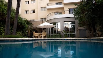 Una piscina con tumbonas y sombrillas, rodeada de vegetación y un edificio de varios pisos con balcones.