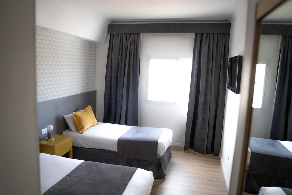 A hotel room with two beds, a bedside table with a lamp, a TV mounted on the wall, and a window with curtains.