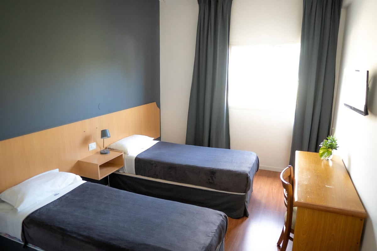 A hotel room with two single beds, a wooden nightstand, a desk with a chair, and a window with curtains.