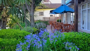 A patio area with wooden tables and chairs, surrounded by lush greenery and flowering plants. - Hotel Rideamus (Villa Gesell)