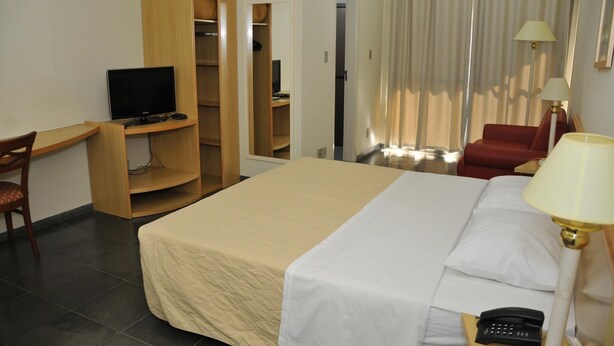 Quarto de hotel com cama, escrivaninha com televisão, cadeira, guarda-roupa e poltrona vermelha.