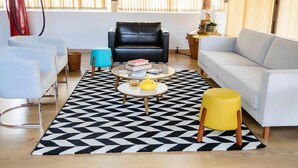 Uma sala de estar com um sofá de couro preto, um sofá branco, uma mesa de centro com livros, um banquinho amarelo e um tapete com padrão preto e branco.