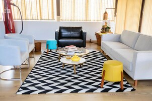 A living room with a black leather sofa, a white sofa, a coffee table with books, a yellow ottoman, and a black and white patterned rug.