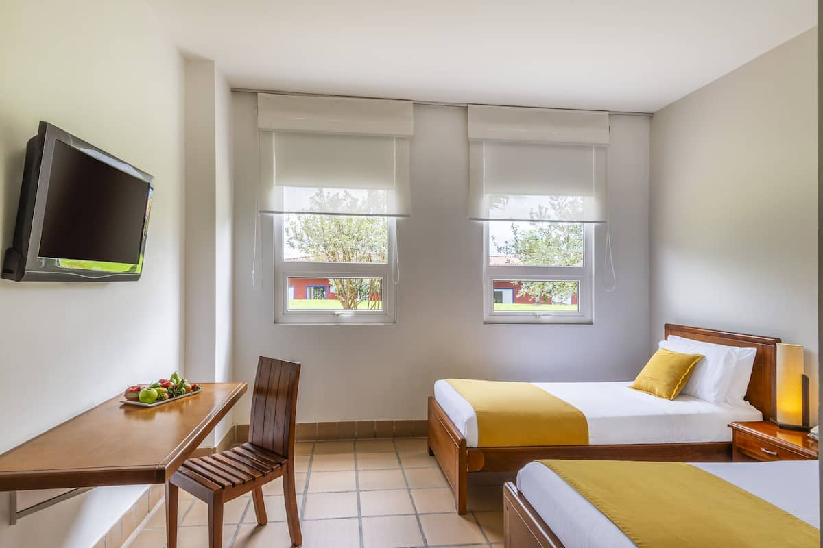 Une chambre d’hôtel avec deux lits, un bureau, une chaise, une télévision et des fenêtres équipées de stores.