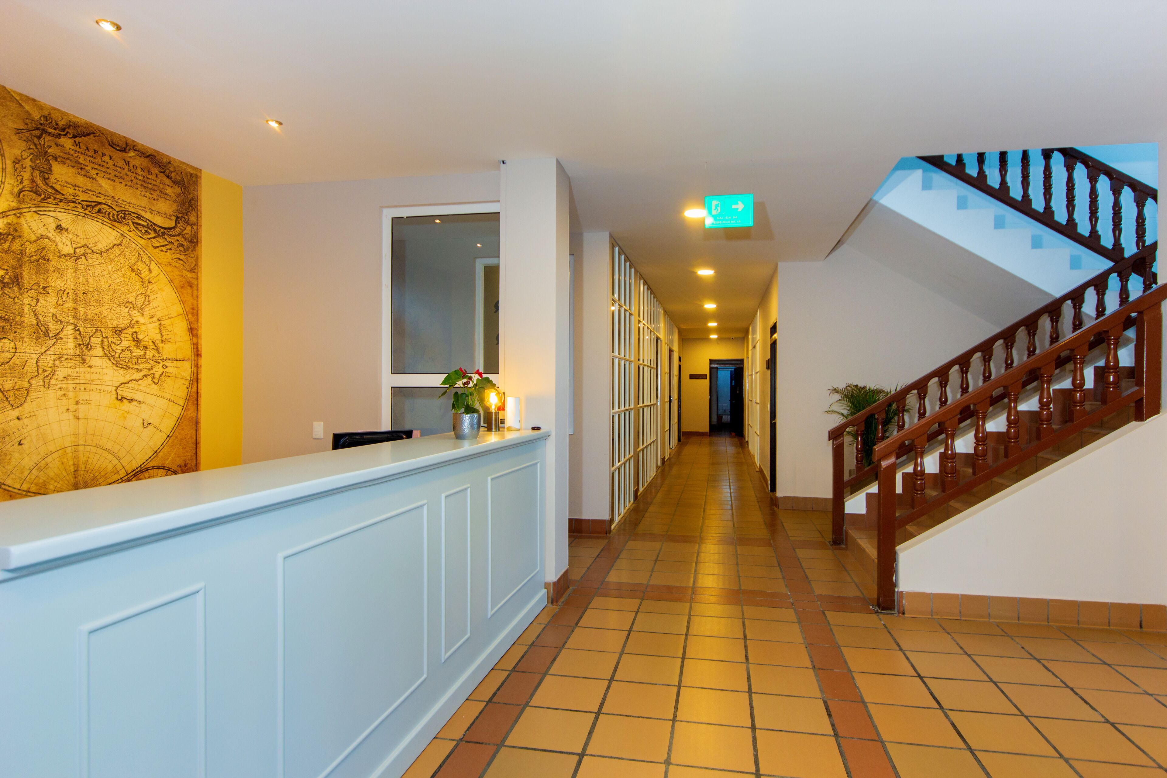 A hotel reception area with a large map on the wall, a reception desk, a staircase, and a hallway leading to other rooms.