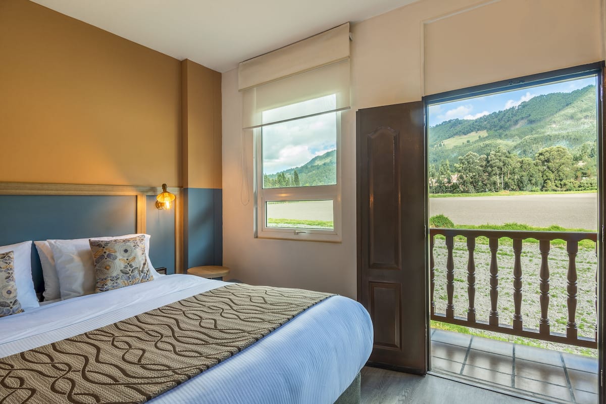 Une chambre d’hôtel avec un grand lit, une table de chevet, une fenêtre donnant sur un paysage verdoyant et un balcon avec une balustrade.