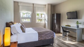A bedroom with a bed, bedside lamp, desk, chair, TV, and windows.