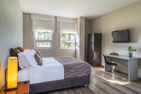 A bedroom with a bed, bedside lamp, desk, chair, TV, and windows.