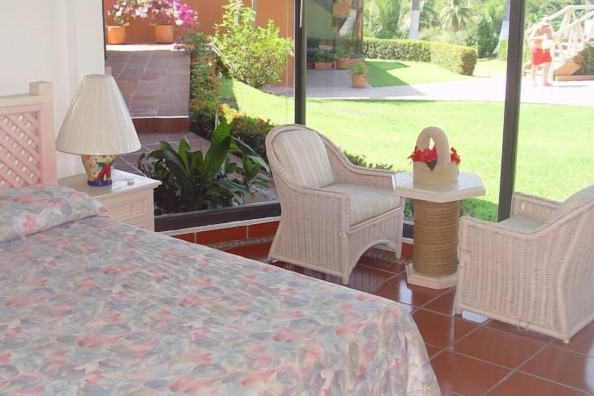 Ein Schlafzimmer mit Bett, Nachttisch, Lampe und Blick auf einen Garten durch große Fenster.