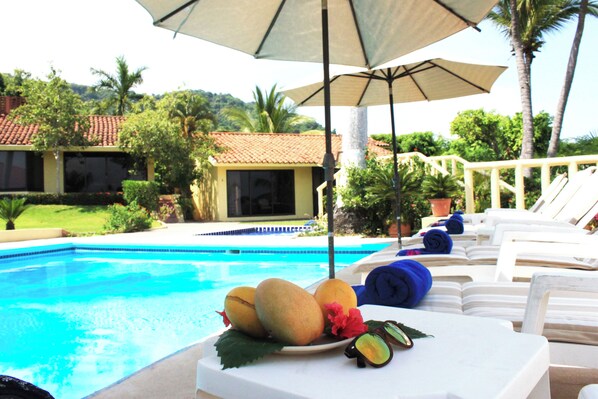 Uma área à beira da piscina com espreguiçadeiras, uma mesa com frutas e um guarda-sol.